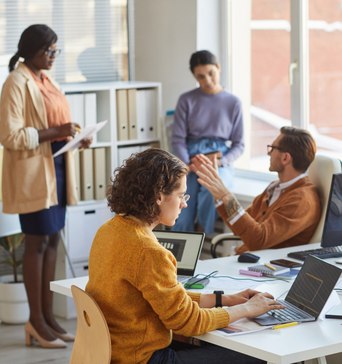 co-workers interacting in a corporate office while one woman sits at a desk working on a laptop